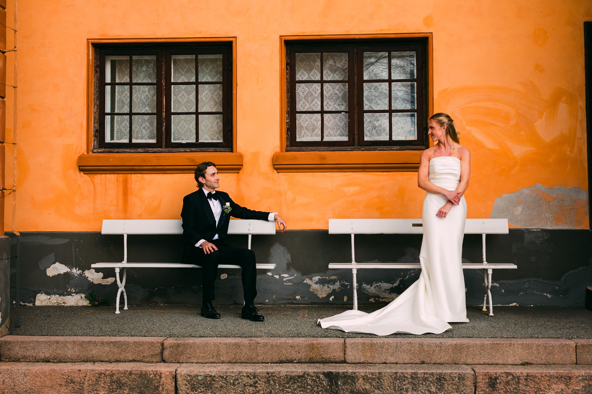 Brudepar foran en gul vegg, brudgom sitter på benk mens brud står